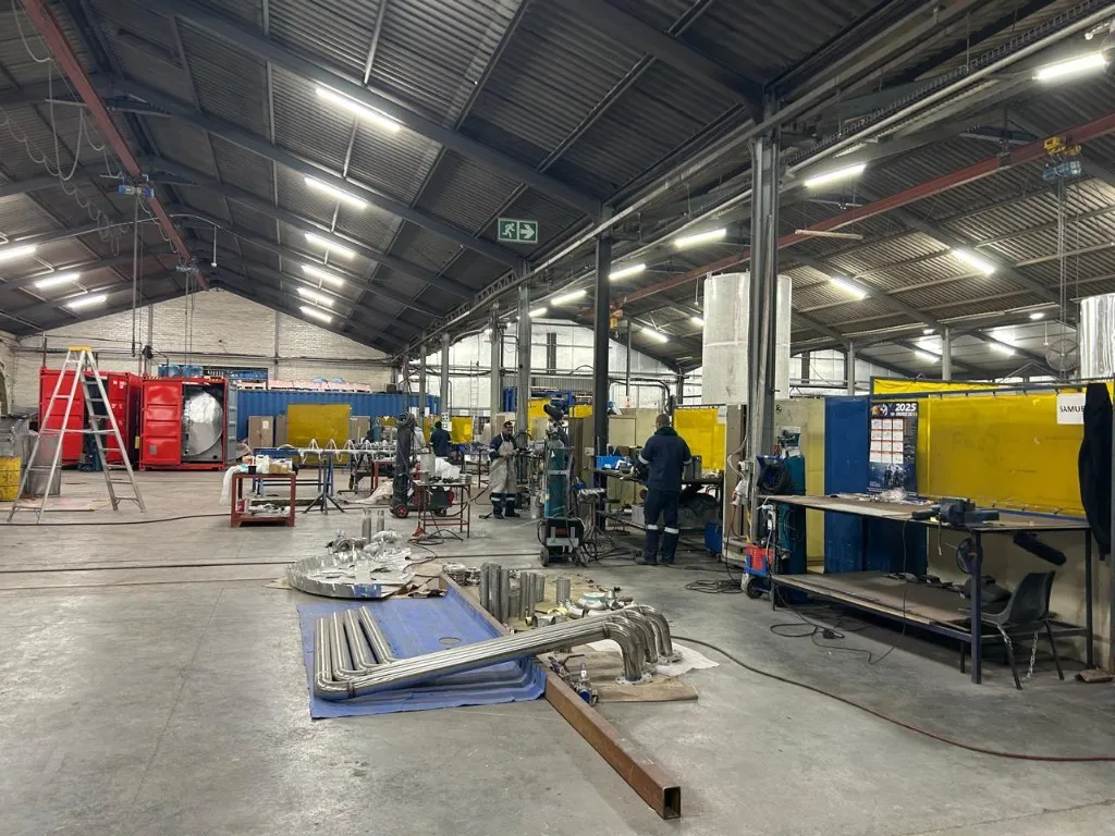 Wide view of a busy metal fabrication workshop with workers, equipment, and stainless steel pipework